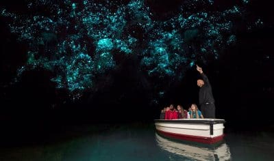 Visitors in a boat marvel at radiant blue glow-worms illuminating New Zealand’s Waitomo Caves, resembling a star-filled night sky.