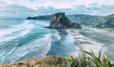 Breathtaking coastal view of Piha Beach featuring black sand, dramatic waves, rocky outcrop, lush green hills, and a moody sky.