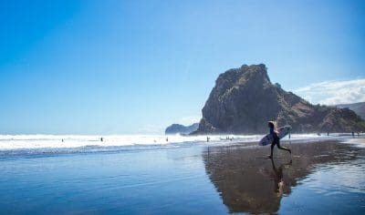 Surfer sprinting across glistening wet sand at Piha Beach, iconic Lion Rock towering behind, under a vibrant blue New Zealand sky.