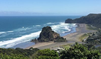 Dramatic rocky outcrop on Piha Beach’s golden sand, lush coastal greenery, vibrant blue sky, and crashing ocean waves in New Zealand.