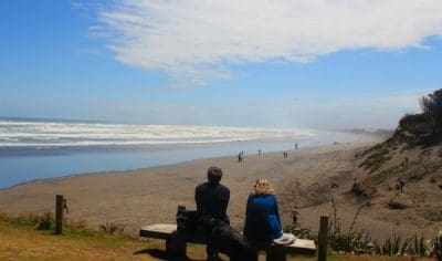 Couple seated on a bench gazes over scenic Muriwai Beach, observing crashing waves and beachgoers under a picturesque cloudy sky.