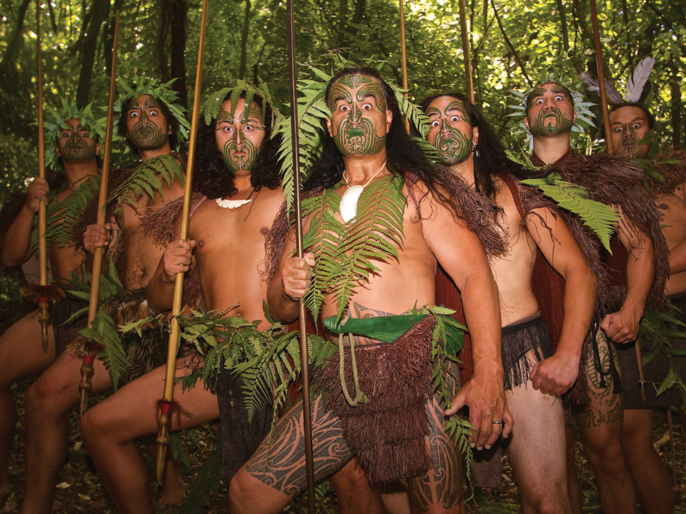 Six Māori warriors with intricate tattoos and spears stand in a lush forest, set to welcome guests to an immersive Māori Dinner Show.