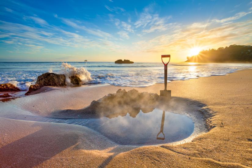 A spade in wet sand by a water-filled hole catches sunrise on Coromandel Beach as ocean waves crash, capturing New Zealand’s beauty.