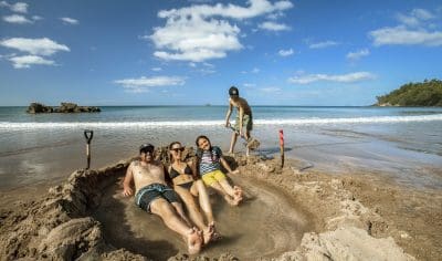 A family enjoys a sandy hot pool at Coromandel Beach as a child digs nearby, framed by ocean waves and clear blue sky in New Zealand.