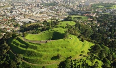 Stunning aerial view of a lush green volcanic hill overlooking Auckland City skyline and sparkling harbour under a vibrant clear sky.