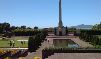Visitors stroll by a towering stone obelisk and tranquil reflecting pool in Auckland City gardens on a clear, sunny day.