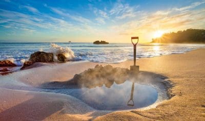 Spectacular sunrise on Coromandel’s sandy beach, spade in steaming Hot Water Beach pool, with ocean waves crashing close by.
