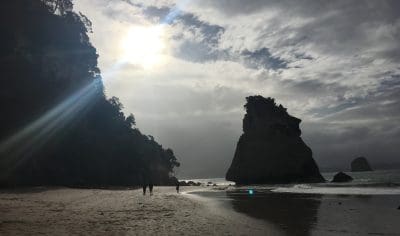 Golden sun rays shine through dramatic clouds above Coromandel beach, highlighting spectacular rock formations along the scenic shore.