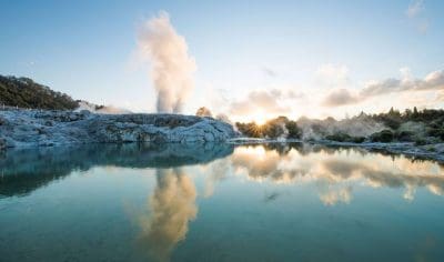 Sunset over Rotorua geothermal pool, with steam rising and mirror-like water reflecting trees and clouds for a serene scenic view.