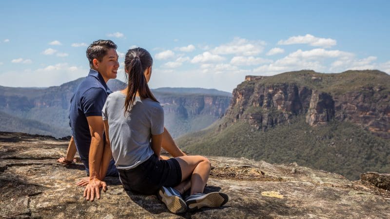 A couple enjoys panoramic mountain views from a rocky cliff on a 1 Day Blue Mountains Sunset Wilderness Tour adventure.