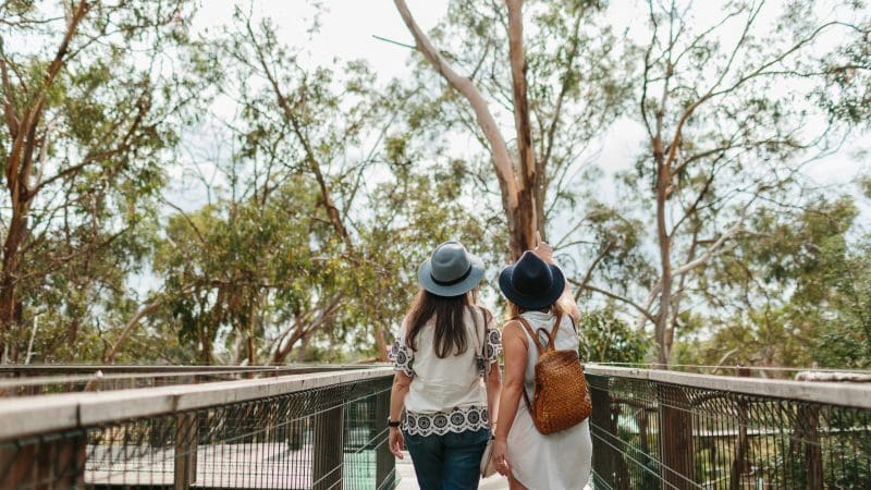 Two women in hats walk hand in hand on a scenic wooden bridge at Phillip Island during the famous Penguin Parade and Koalas tour.