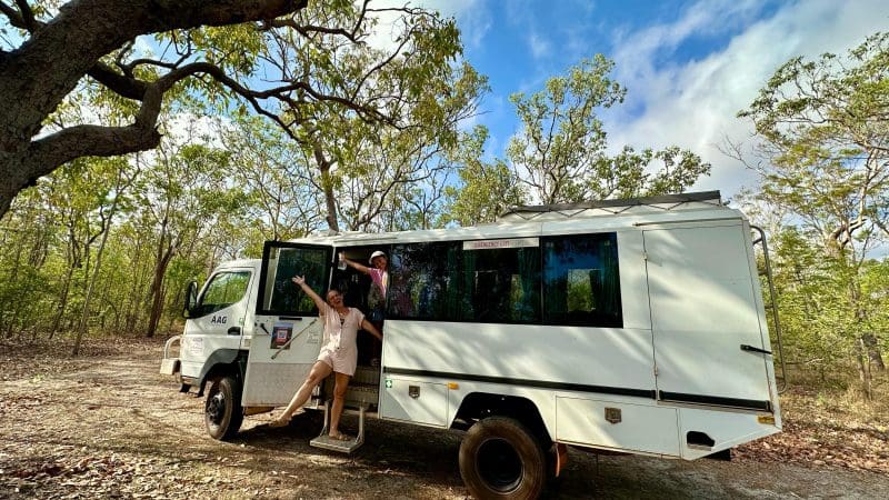Two happy travellers smile and wave from the open door of a white camper lorry during their Kakadu National Park adventure tour.