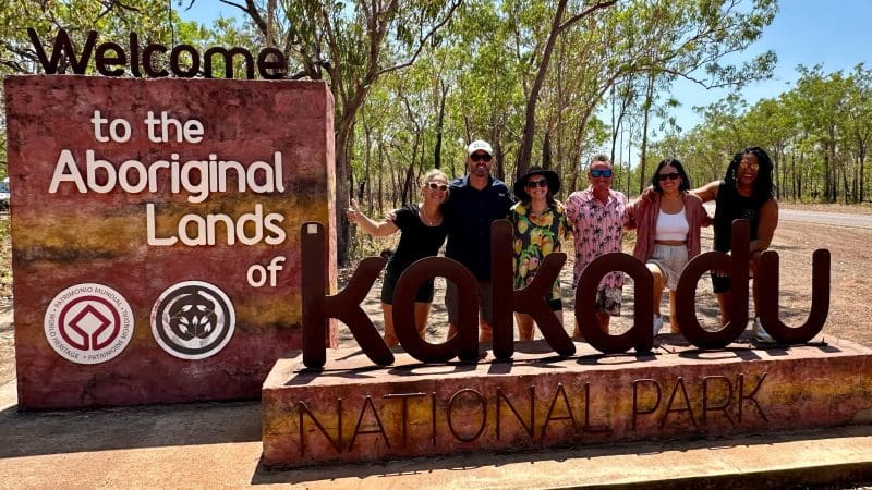 Group of six happy campers by the Welcome to Kakadu sign during a 3 Day Kakadu Adventure, June–Sept, in bright, wooded outback.