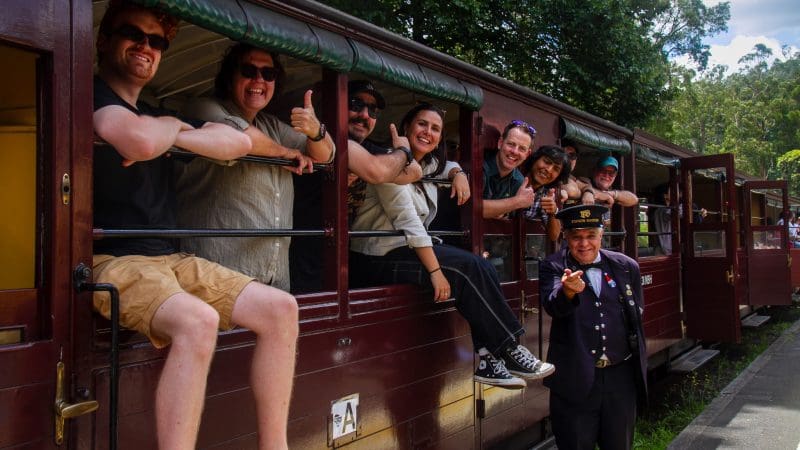 Happy group leans from a Puffing Billy Steam Train carriage, posing with friendly guard on the platform for memorable tour photo.