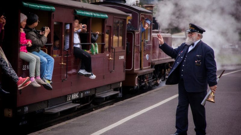 Train guard waving to excited children on Puffing Billy Steam Train, holding brass horn, with clouds of steam billowing in the background.