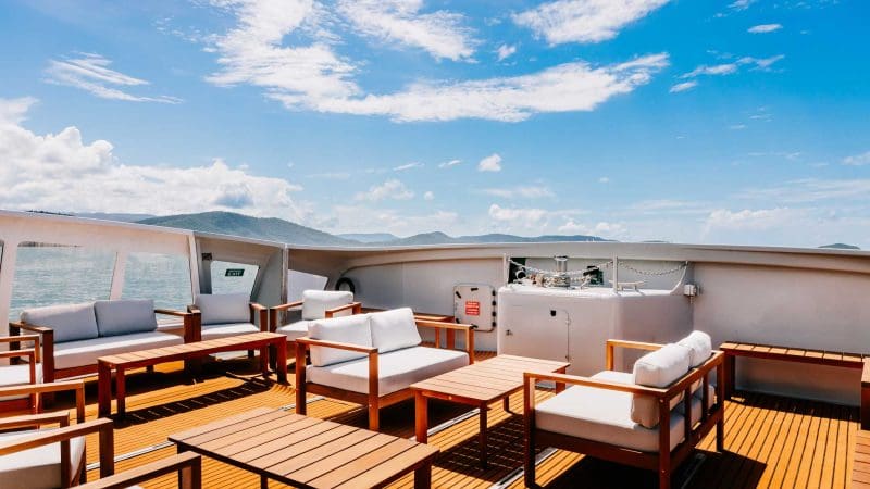 Luxurious yacht deck with plush seating, stylish wooden tables, and panoramic Whitehaven Beach views—sun-soaked relaxation all day.