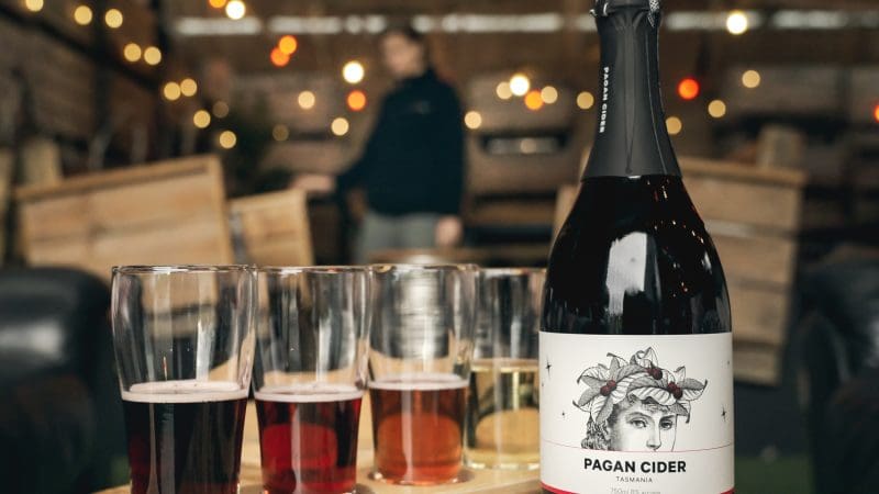 Pagan Cider bottle and four glasses on a table, ideal for relaxing after a Huon Valley Safaris Private Day Tour experience.