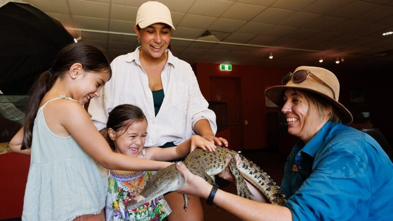 Two children and two adults happily interact with a Crocosaurus Cove keeper, touching a small crocodile during an indoor wildlife tour.