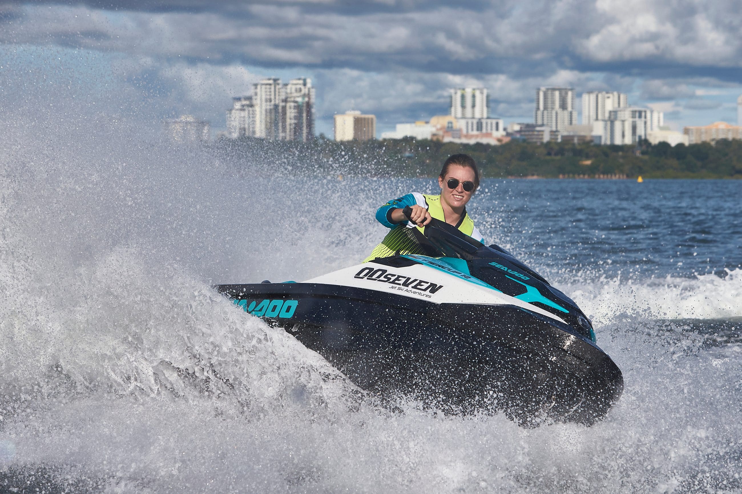 Jet ski rider speeding by Thunderball Shipwreck, sparkling blue waters, city skyline, lush trees; thrilling Bahamas adventure scene.
