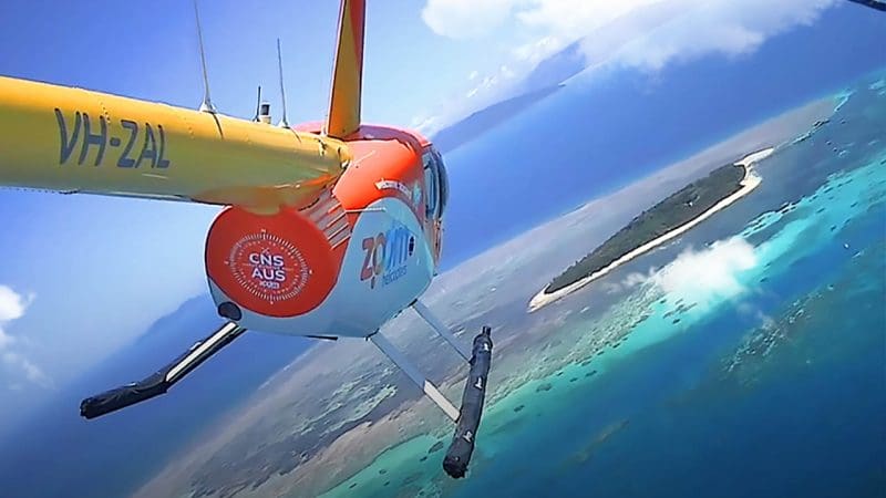 Scenic helicopter soars above vibrant turquoise waters and lush island on a 40 Minute Outer Reef Odyssey under blue sky with clouds.