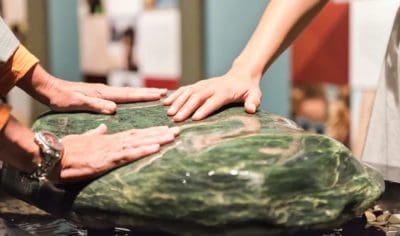 Three hands touch a polished green stone at New Zealand’s historic Waitangi Treaty Grounds, symbolising unity and cultural heritage.