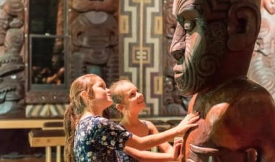 Two girls smile and touch a massive Māori statue at the intricately decorated Waitangi Treaty Grounds, New Zealand’s historic site.