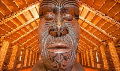 Detailed close-up of intricately carved wooden Māori statue at Waitangi Treaty Grounds, inside meeting house with ornate ceiling décor.