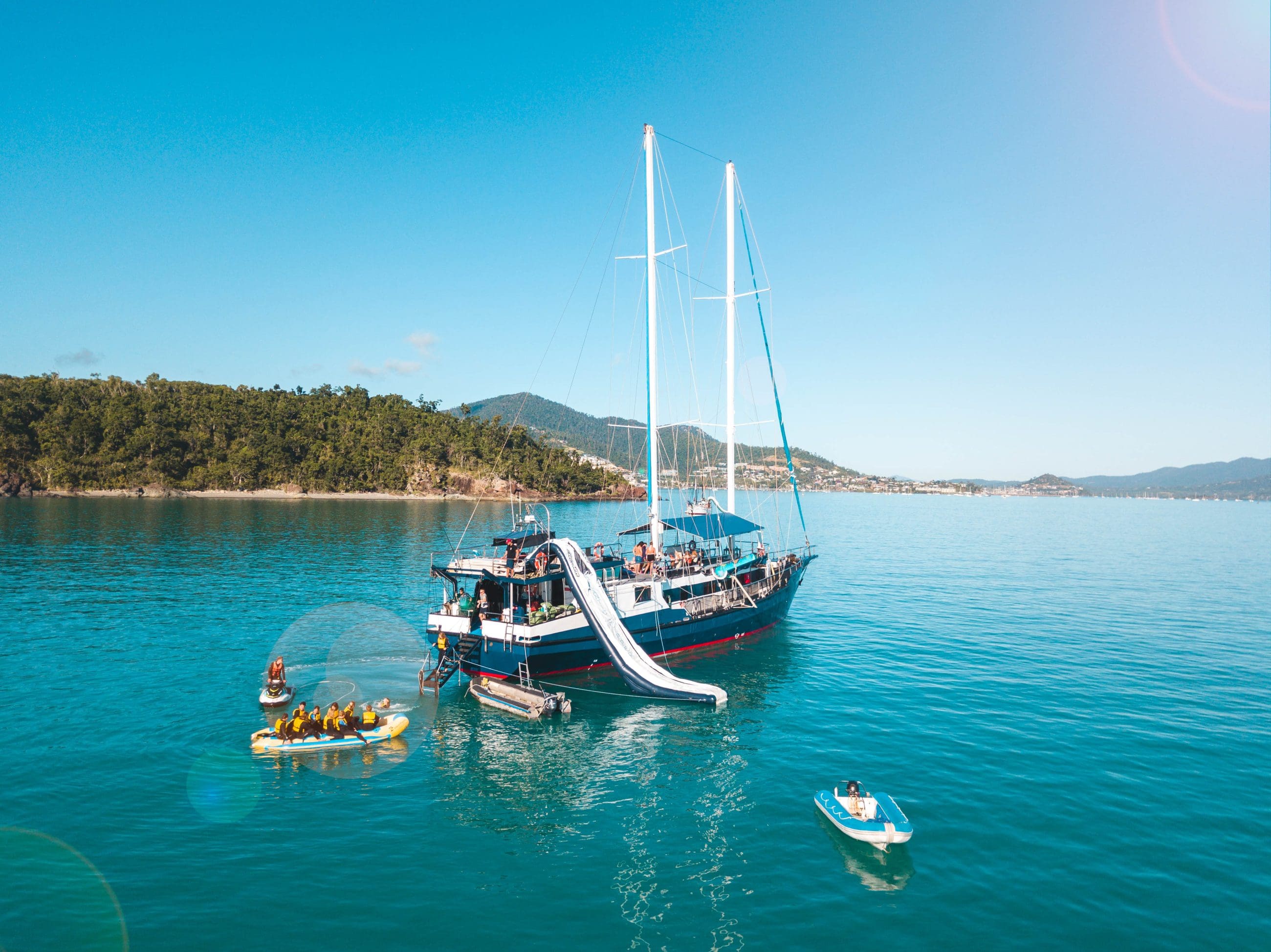 Experience The Great Escape Australia: 28-Day Semi-Guided yacht with water slides, anchored in crystal-clear waters amid small boats.