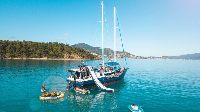 Experience The Great Escape Australia: 28-Day Semi-Guided yacht with water slides, anchored in crystal-clear waters amid small boats.