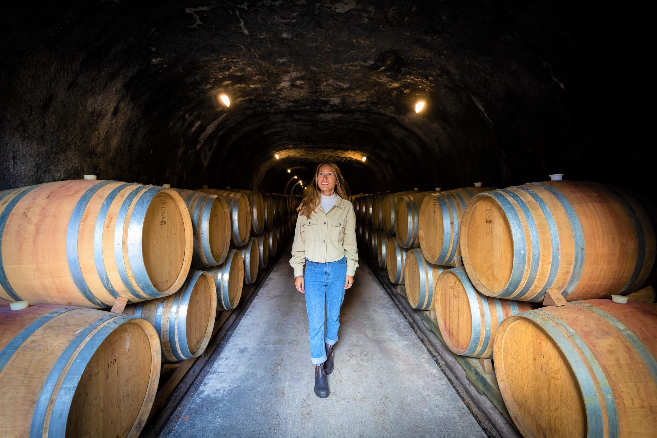A woman explores a dimly lit wine cellar tunnel, walking between stacked barrels on a luxurious gourmet wine tasting tour experience.