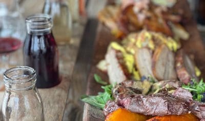 Gourmet wooden platter with grilled meats, roasted squash, artisan sandwiches on rustic table; empty glass jars accent the scene.