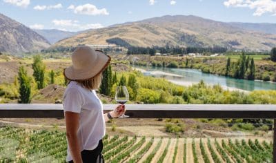 A woman in a sunhat savours premium wine on a luxury boutique wine tour, overlooking scenic vineyards, majestic mountains, and winding river.