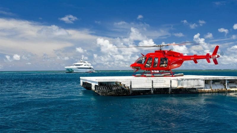 Red helicopter parked on dock beside luxury white yacht for Get High Package Full Day Reef Cruise and Helicopter Flight adventure.