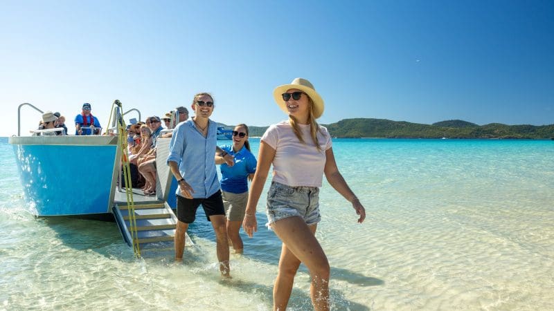 Guests smiling and disembarking from a Purely Whitehaven Day Tour boat, with turquoise waters and lush Whitsunday island scenery behind.