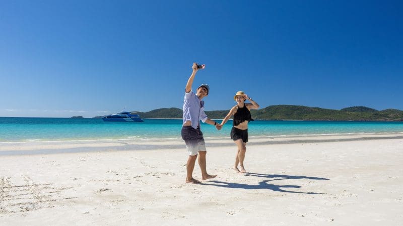 Smiling couple holding hands and taking a selfie on a Purely Whitehaven Day Tour, framed by turquoise sea and lush green hills.