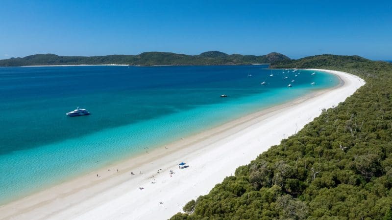 Stunning aerial shot of Purely Whitehaven Day Tour: pristine white sands, crystal-clear turquoise waters, boats, lush forest, blue sky.