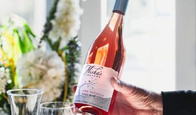 A hand displays Misha’s Vineyard The Soloist Pinot Rosé wine bottle by a glass and fresh flowers, illuminated in vibrant sunlight.