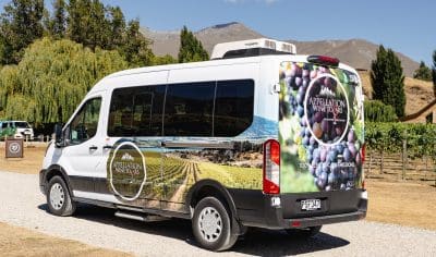 Boutique Appellation Wine Tours van featuring branded graphics, parked on a gravel road beside lush trees with scenic mountains behind.