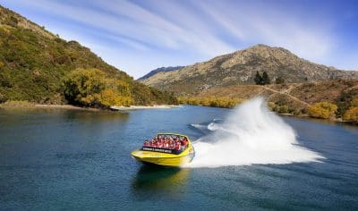 Thrilling yellow jet boat speeds down a scenic river, splashing water with passengers aboard, framed by lush hills and clear blue sky.