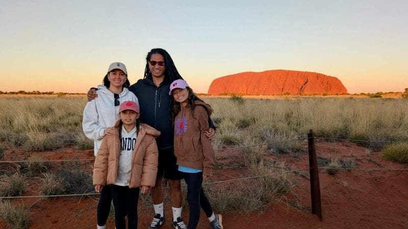Family of four at Uluru Red Centre at sunset, standing on iconic red earth with golden grasses and clear blue skies in the Australian Outback.