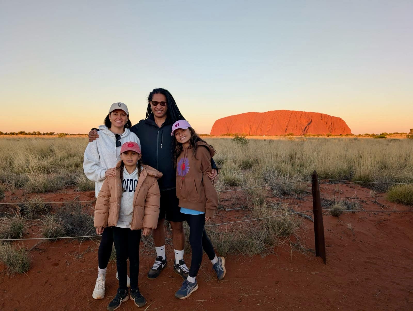 Family of four at Uluru Red Centre at sunset, standing on iconic red earth with golden grasses and clear blue skies in the Australian Outback.
