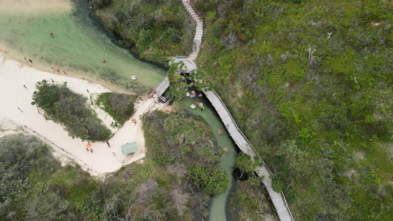 Stunning aerial shot of winding wooden boardwalk on Kgari 2 Day Tour, Rainbow Beach; swimmers relax in creek surrounded by lush trees.