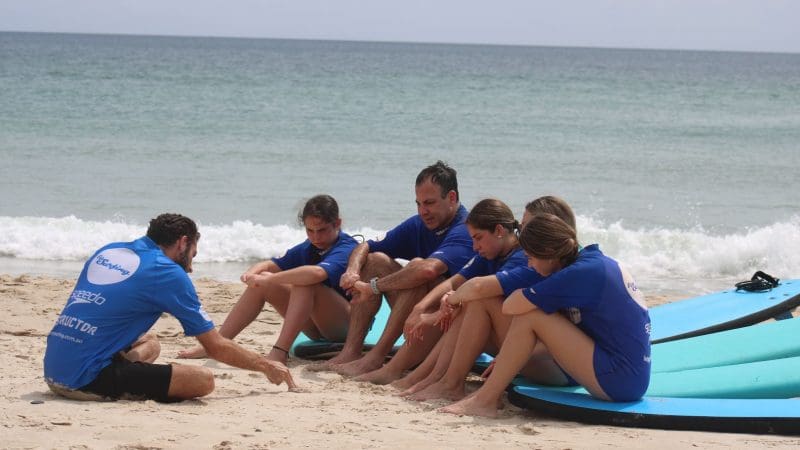 Surf instructor teaching beginners on Byron Bay beach during the Learn to Surf Half-Day Tour, group listens on sandy shoreline.