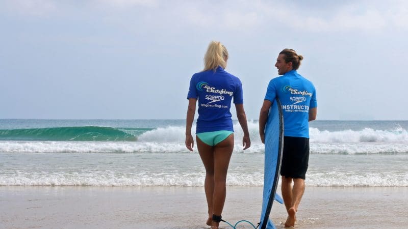 Two surfers in blue shirts stand on Byron Bay beach, prepared for their Learn to Surf Half Day Tour—ultimate surf lesson experience.