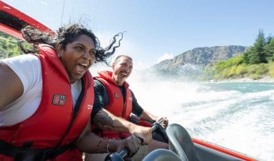 Two people in red life jackets enjoy a thrilling Shotover Jet ride, splashing water with stunning Queenstown mountains in the background.