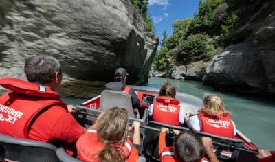 Thrill-seekers in red life jackets speed on a Shotover Jet through Queenstown’s stunning blue gorge under a bright, sunny sky.