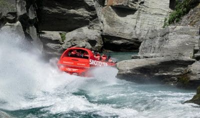 Experience the thrill as a red Shotover Jet surges through Queenstown’s rocky river rapids, helmeted riders gripping tightly on board.