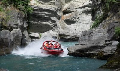 Experience the thrill as a red Shotover Jet boat races through Queenstown’s narrow rocky canyon, skimming blue waters and splashing.