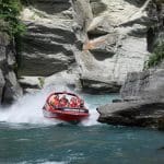 A bright red Shotover Jet boat races through a scenic rocky canyon over crystal-clear water, splashing mist as excited riders hold on.
