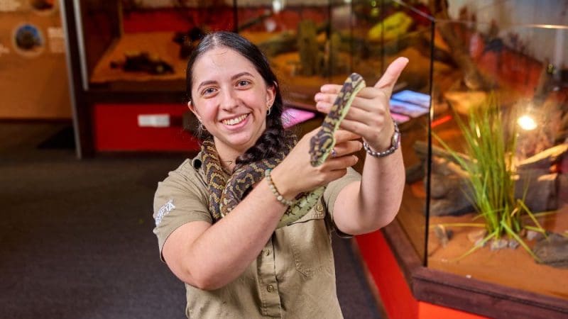 Smiling woman in khaki shirt holding a python at Cairns City Sights reptile exhibit, with glass animal enclosures in the background.